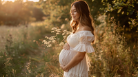 Femme enceinte souriante touche son visage lumineux, illustrant un joli teint pendant la grossesse dans un cadre lumineux.