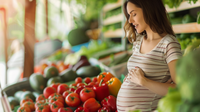 Une femme enceinte souriante prépare des aliments sains et colorés sur une table dans une cuisine lumineuse.